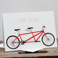A greetings card with a drawing of two red bicycles and the text 'MADE FOR TWO' on it, placed on a wooden surface.