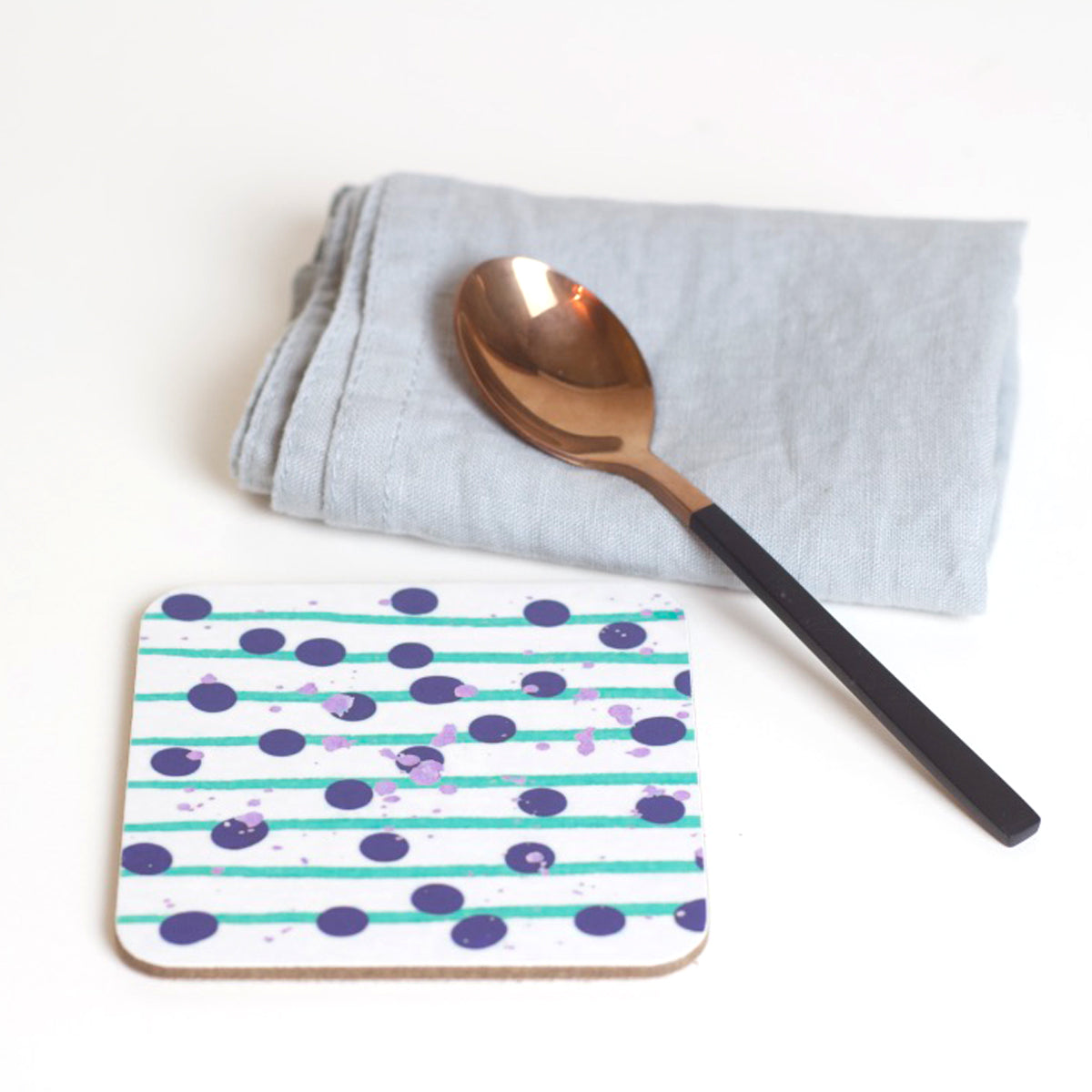 A square coaster with a geometric pattern of green lines and blue dots, accompanied by a copper-colored spoon and a grey napkin.