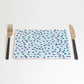 A rectangular placemat with a green and blue dot pattern, featuring a fork on the left side and a knife on the right side, placed on a white background.