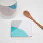A square coaster with teal and grey color dip design, placed next to a bone china mug and a wooden spoon.
