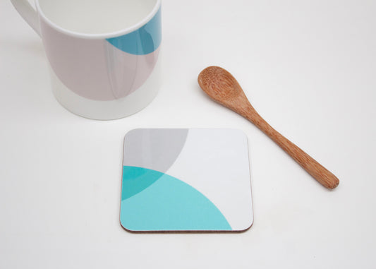 A square coaster with teal and grey color dip design, placed next to a bone china mug and a wooden spoon.