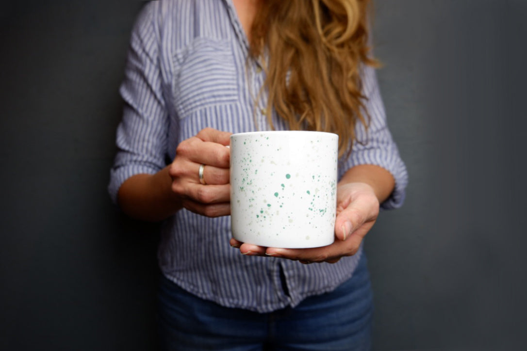 A person holding a white bone china mug with teal and grey speckled pattern.