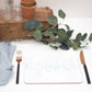 A splatter placemat in teal color with a speckled pattern, placed on a white surface with a fork, knife, and a small round table in the background with a potted plant and some decorative leaves.