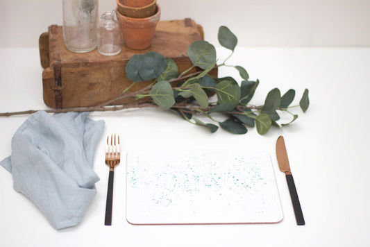 A splatter placemat in teal color with a speckled pattern, placed on a white surface with a fork, knife, and a small round table in the background with a potted plant and some decorative leaves.