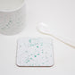A white coaster with teal splatter design, placed on a white surface next to a white mug and a white spoon.