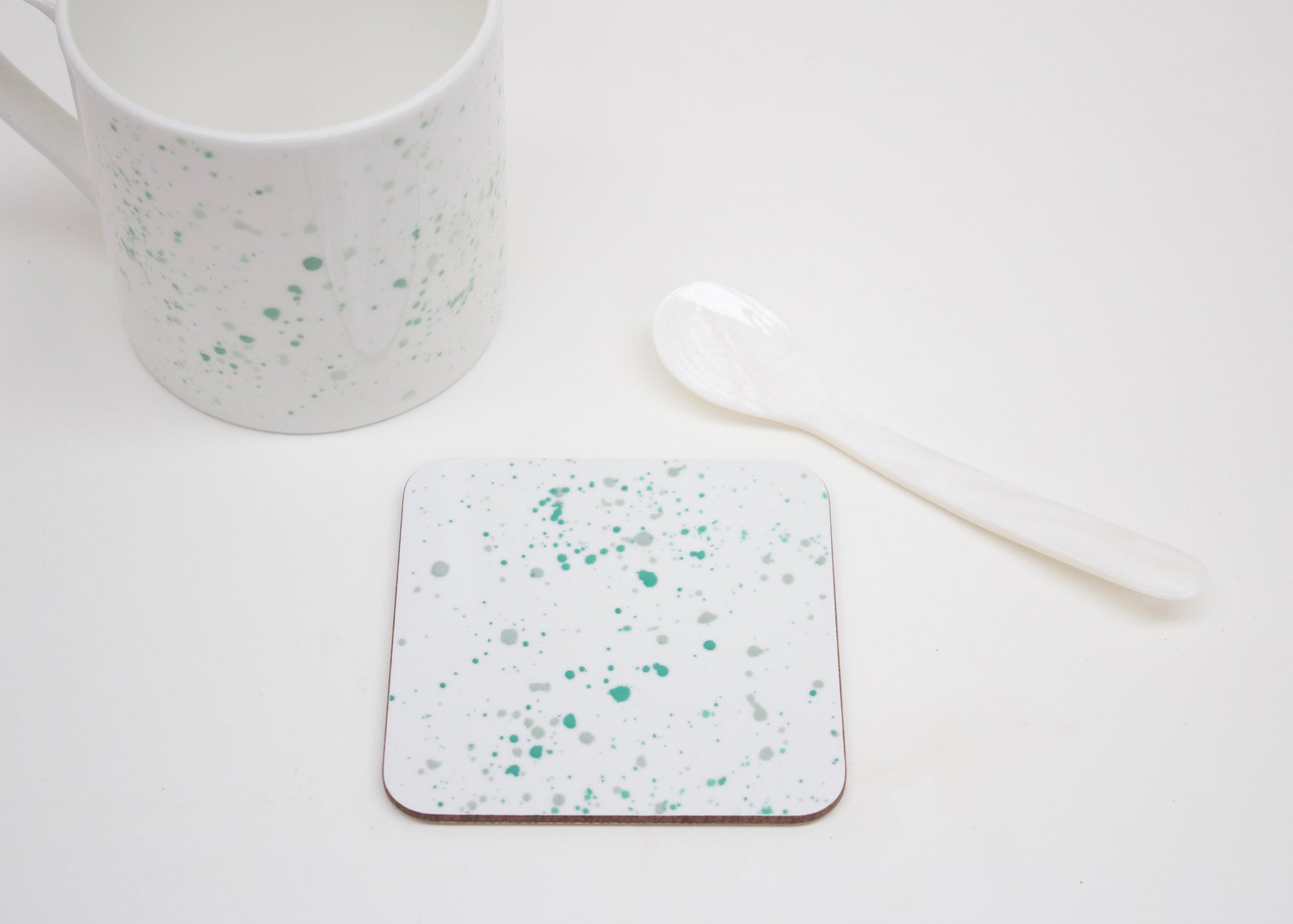 A white coaster with teal splatter design, placed on a white surface next to a white mug and a white spoon.