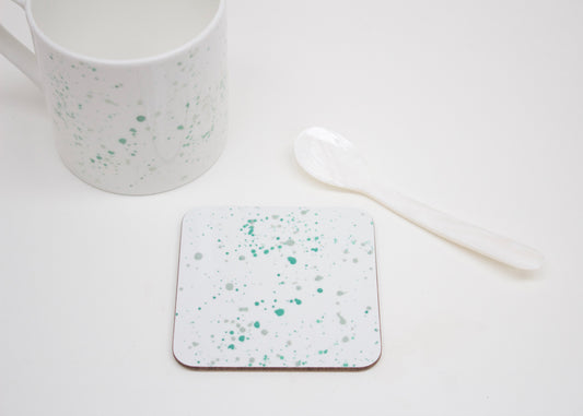 A white coaster with teal splatter design, placed on a white surface next to a white mug and a white spoon.