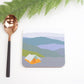 A square coaster with a camping tent design in the Picos mountains, accompanied by a copper-colored spoon and green foliage in the background.