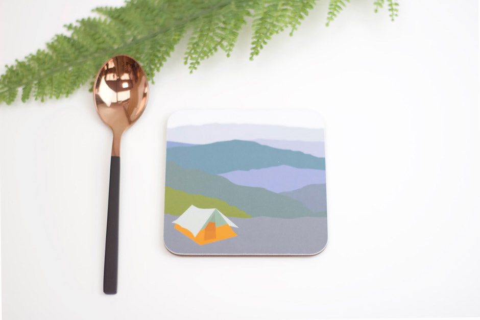 A square coaster with a camping tent design in the Picos mountains, accompanied by a copper-colored spoon and green foliage in the background.