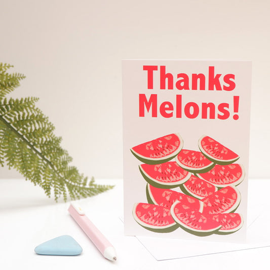 A greeting card with the text 'Thanks Melons!' printed on it, featuring a design of stylized melons. The card is displayed upright with a pink pen, and a decorative leaf in the background.