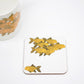 A square coaster with a hand-drawn goldfish design on a white background, placed on a white surface next to a white cup with a similar design.