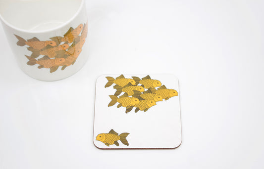 A square coaster with a hand-drawn goldfish design on a white background, placed on a white surface next to a white cup with a similar design.