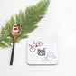 A coaster with drawings of three bikes on it, placed on a white surface next to a green fern leaf and a spoon with a bicycle design.
