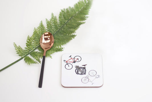 A coaster with drawings of three bikes on it, placed on a white surface next to a green fern leaf and a spoon with a bicycle design.