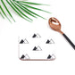 A white coaster with black symbol designs, a bronze-colored spoon, and green palm leaves in the background.