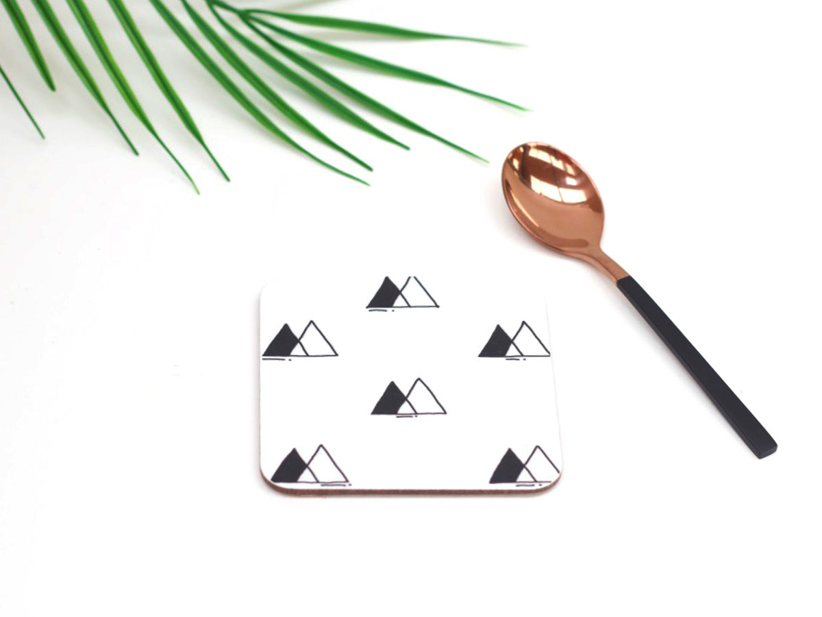 A white coaster with black symbol designs, a bronze-colored spoon, and green palm leaves in the background.