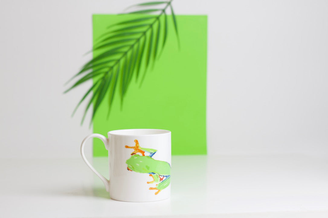A green bone china mug with a design of a tree frog climbing up the mug, placed on a white surface with a palm leaf in the background.