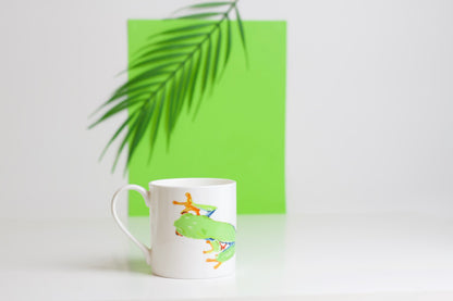 A green bone china mug with a design of a tree frog climbing up the mug, placed on a white surface with a palm leaf in the background.