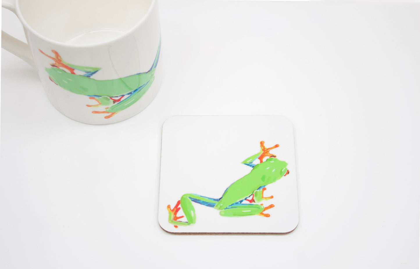 A square coaster with a hand-drawn tree frog design, part of the Jungle Collection, placed on a white surface next to a white mug with a similar design.