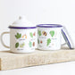 Two enamel mugs with tree leaf illustrations, with a lid, on a wooden board. 