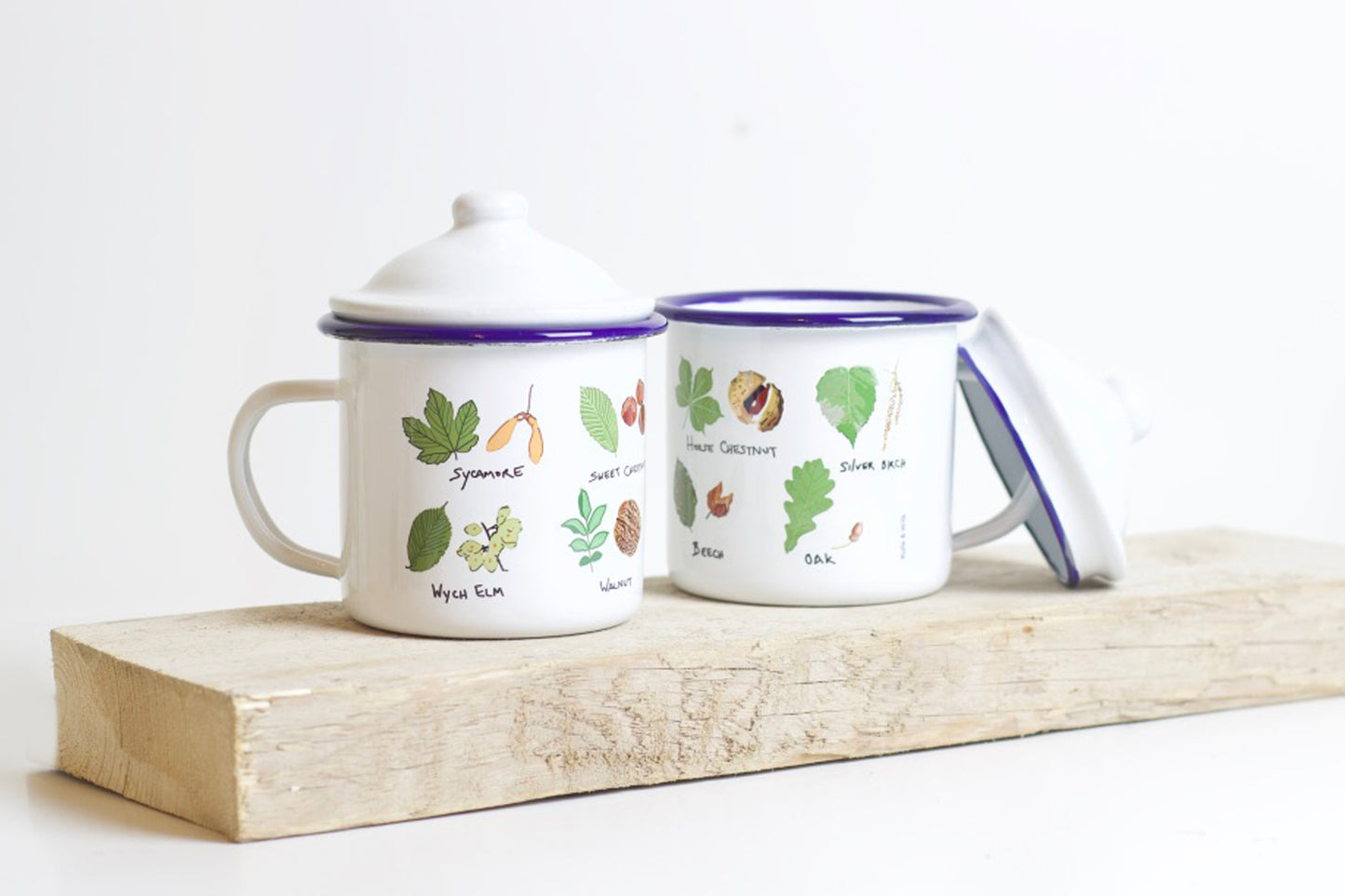 Two enamel mugs with tree leaf illustrations, with a lid, on a wooden board. 