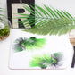 A placemat featuring a vibrant green tropical cabbage leaf design, displayed on a table with a piece of bamboo cutlery, a glass vase with a palm plant, and other decorative greenery in the background.