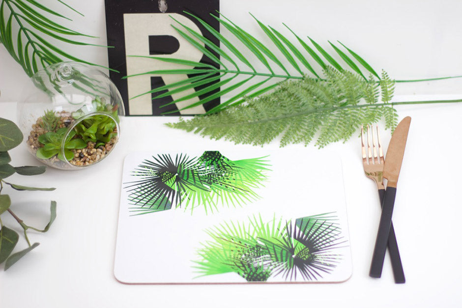 A placemat featuring a vibrant green tropical cabbage leaf design, displayed on a table with a piece of bamboo cutlery, a glass vase with a palm plant, and other decorative greenery in the background.