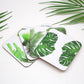 A set of four white coasters with green tropical leaf designs, placed on a surface with more leaves and a palm plant in the background.