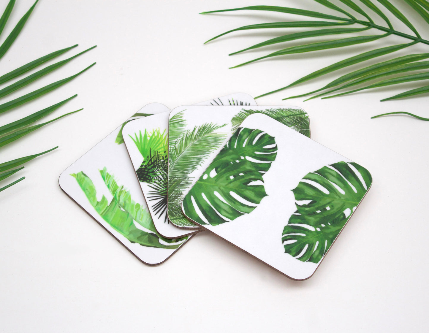 A set of four white coasters with green tropical leaf designs, placed on a surface with more leaves and a palm plant in the background.