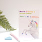 A greeting card with an illustration of a unicorn and colorful text on the front, placed on a table with a plant in the background.