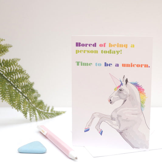 A greeting card with an illustration of a unicorn and colorful text on the front, placed on a table with a plant in the background.