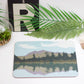 A rectangular placemat with a lake and trees design, placed on a white surface with a fork and knife to the side, beside a potted plant and decorative items.