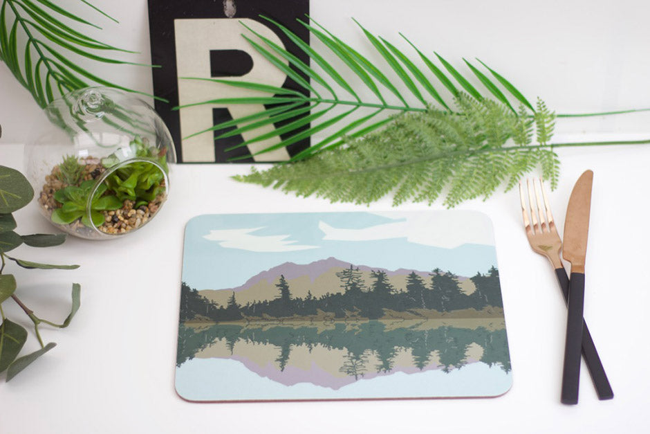 A rectangular placemat with a lake and trees design, placed on a white surface with a fork and knife to the side, beside a potted plant and decorative items.