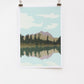 A digital print of a lake scene with mountains in the background, hanging on a wall with two clip bookmarks on the sides.