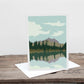 A greetings card with a landscape image of a lake and mountains on it, placed on a wooden surface.