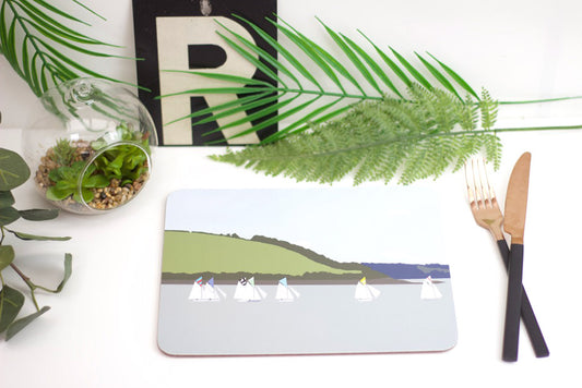 A placemat with a boat design, laid out on a table with cutlery and a plant in the background.
