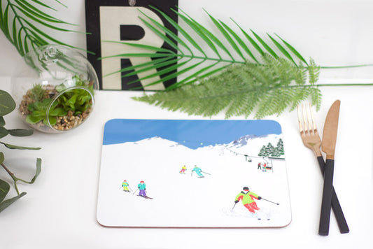 A placemat with a ski-themed design, featuring skiers on a snowy mountain, placed on a dining table with cutlery and a plant in the background.