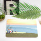 A placemat with a beach and surf design, placed on a table with a fork, knife, and a plant in the background.