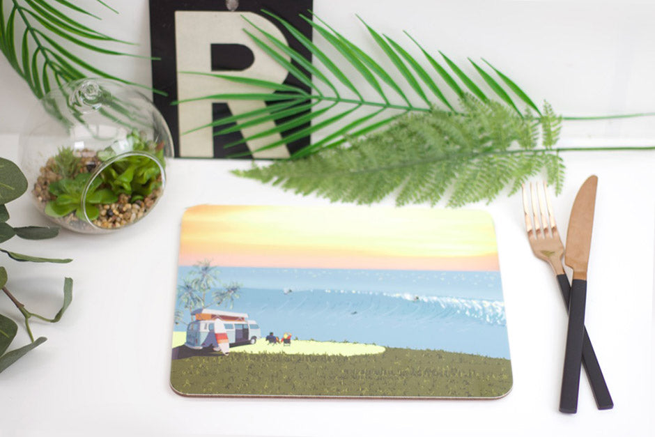 A placemat with a beach and surf design, placed on a table with a fork, knife, and a plant in the background.