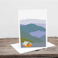 A greetings card with a mountain range and a tent design, placed on a wooden surface.