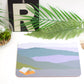 A rectangular placemat with a coastal scene design, placed on a table with a fork and knife on top, beside a potted plant and decorative items.