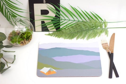 A rectangular placemat with a coastal scene design, placed on a table with a fork and knife on top, beside a potted plant and decorative items.