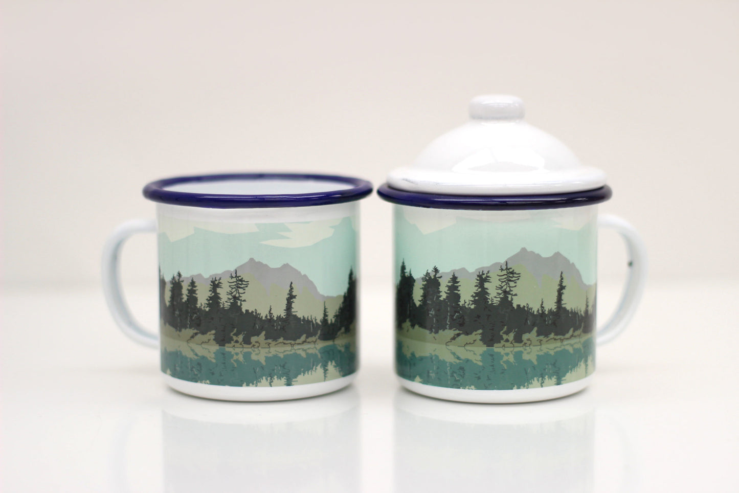 Two enamel mugs with lids, featuring a hand-illustrated design of a lake and forest scene.
