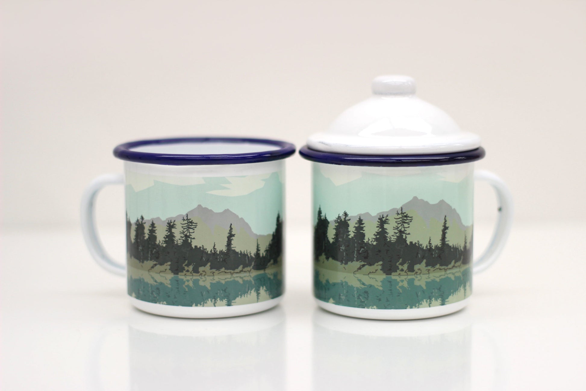 Two enamel mugs with lids, featuring a hand-illustrated design of a lake and forest scene.