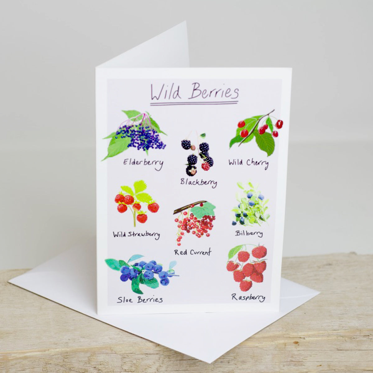 A greetings card with illustrations of various wild berries, including blueberries, blackberries, and raspberries, with the title 'Wild Berries' at the top. Accompanied by a white envelope, placed on a wooden surface.