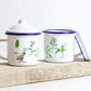 Two white enamel mugs with green and gray illustrations of wild plants, placed on a wooden board. One mug has a lid with a handle.