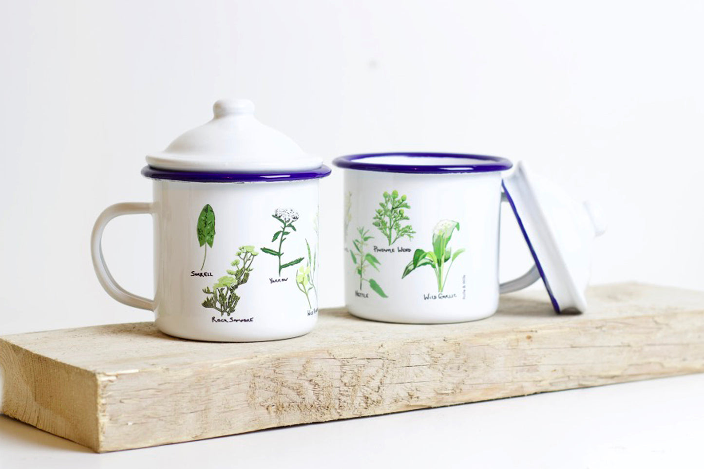 Two white enamel mugs with green and gray illustrations of wild plants, placed on a wooden board. One mug has a lid with a handle.