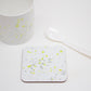 A white coaster with yellow splatter design, placed on a white surface with a cup and a white spoon next to it.