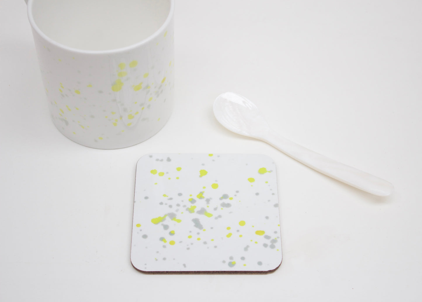 A white coaster with yellow splatter design, placed on a white surface with a cup and a white spoon next to it.