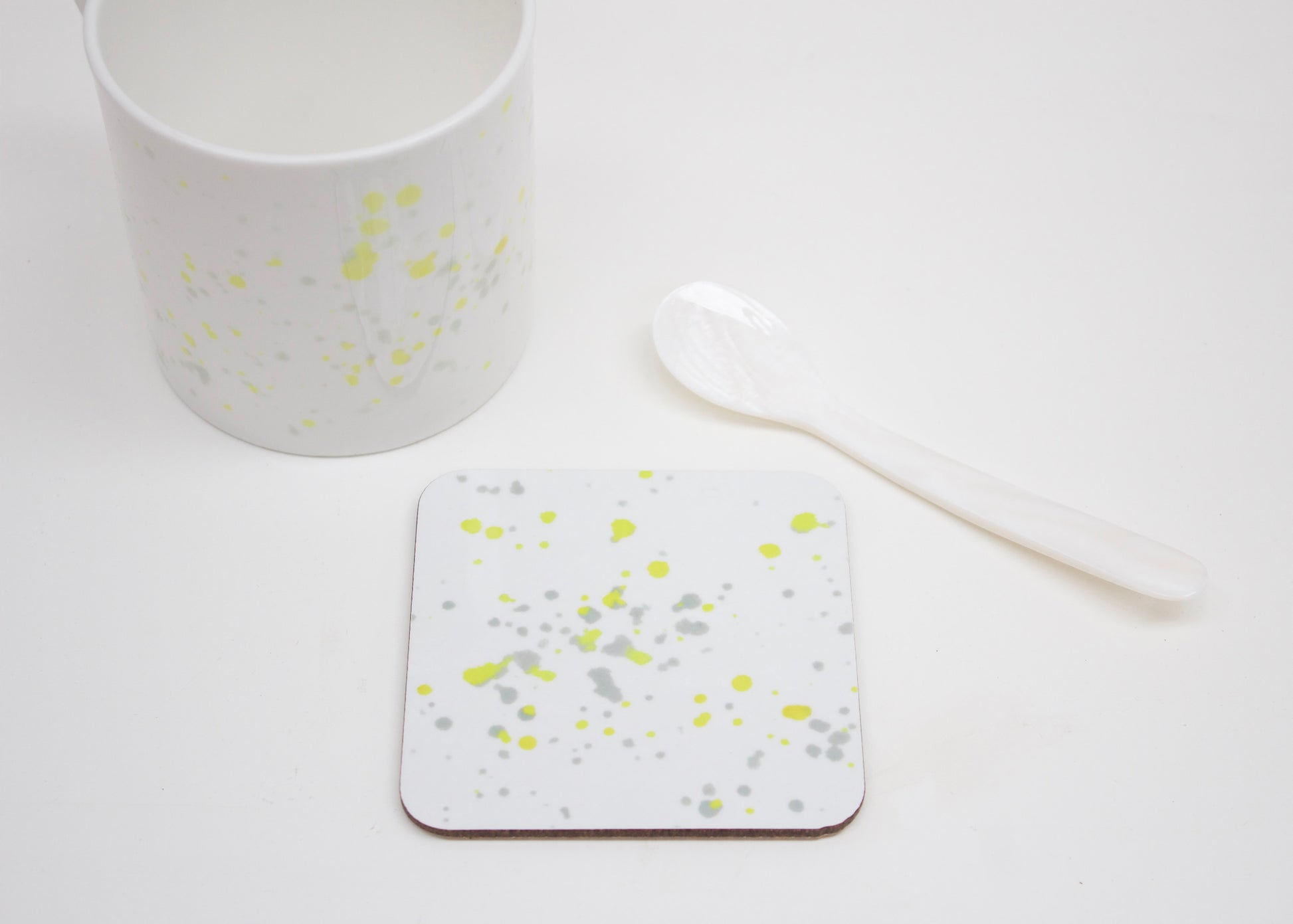 A white coaster with yellow splatter design, placed on a white surface with a cup and a white spoon next to it.